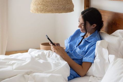 Woman relaxing in blue pajamas smiling and using smartphone in cozy minimal bedroom