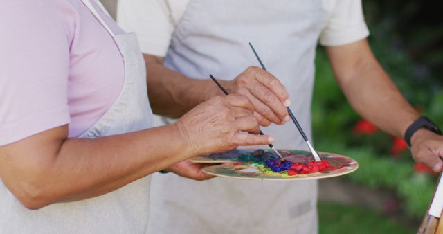 Two seniors enjoy an outdoor acrylic painting activity, collaborating on a colorful palette, which can be used for content related to senior hobbies, community activities, creative workshops, and promoting active lifestyles among the elderly.