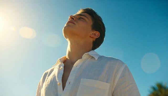 Young man tilting head toward sun wearing white linen shirt sunny coastal day lens flare