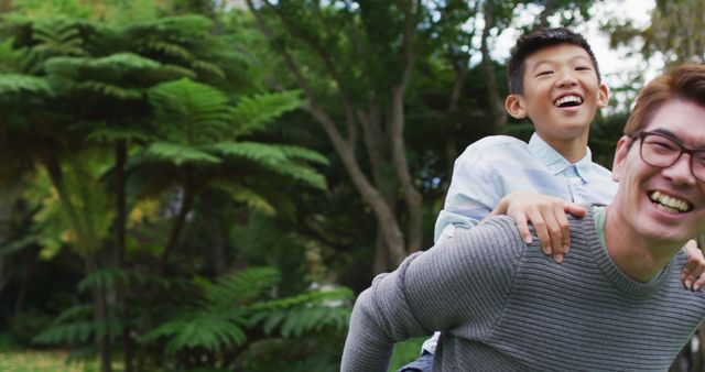 Father and son laughing while enjoying a piggyback ride in a lush park filled with greenery and tall trees. Perfect for use in advertisements for family activities, parenting blogs, outdoor lifestyle campaigns, and family-oriented publications.