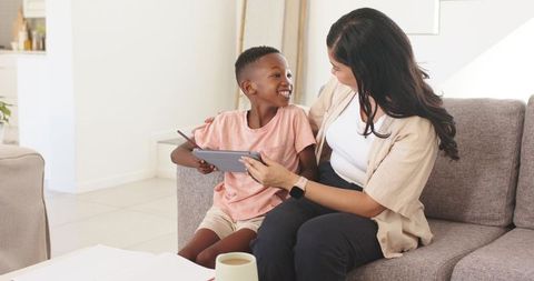 Mother and son sharing tablet on couch, smiling and learning together in cozy sunlit home