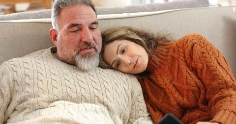 Middle-aged couple cuddling on couch wearing cable knit sweaters sharing cozy moment