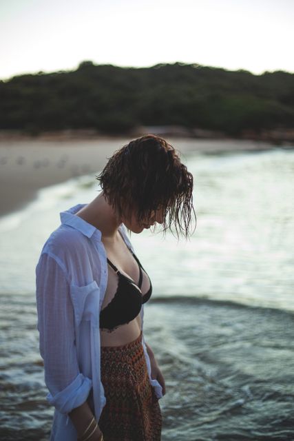 This image depicts a woman in casual beachwear enjoying a tranquil moment by the ocean. Her wet hair indicates she may have just taken a swim. This stock photo can be used for wellness blogs, travel destinations promotions, advertisements for swimwear, or any content focusing on relaxation and nature appreciation.