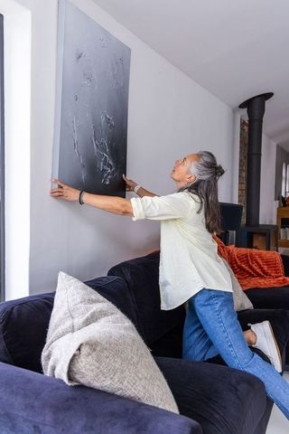 Senior woman hanging dark abstract canvas above navy sofa in modern sunlit living room