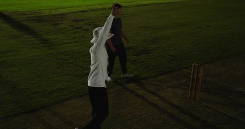 Cricket umpire raising arm during night match on grass pitch with stumps and shadows