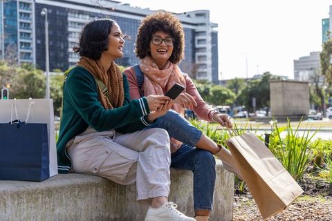 Multiracial young women chatting on urban bench holding shopping bags and smartphone
