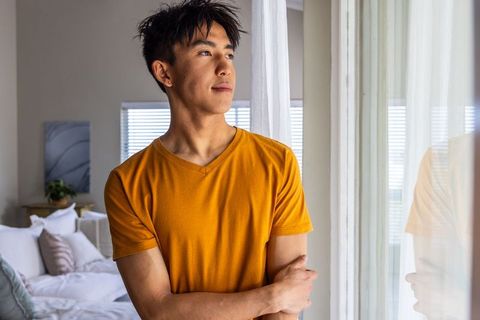 Thoughtful asian man standing by window with sheer curtains in bright modern bedroom