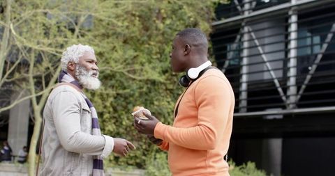 Older African American Man Talking with Young Man Holding Pastry and Headphones in Urban Plaza