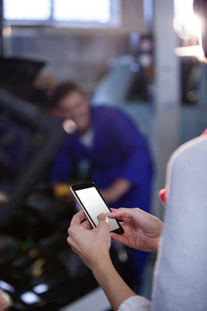 Customer using mobile phone while mechanic works on car in background. Ideal for illustrating automotive services, customer communication, and technology use in everyday life.