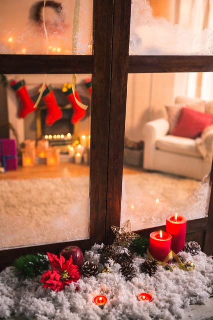 Warm and inviting Christmas scene viewed through a frosted window. Red candles, poinsettia, pine cones, and festive decorations on a bed of fake snow create a cozy holiday atmosphere. Ideal for holiday greeting cards, festive advertisements, and seasonal blog posts.
