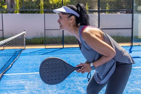 African american woman playing padel on blue court smiling and leaning ready at net