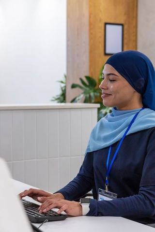 Smiling receptionist wearing headscarf typing at modern front desk with smartwatch, plants