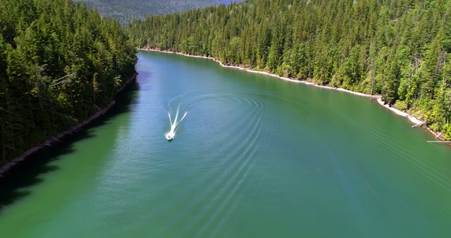 A boat creates ripples as it cruises through the emerald waters of a forest-lined lake, with copy space. The aerial perspective captures the serene beauty of the natural landscape and the leisure activity.