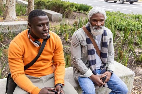 African american men sitting on bench holding smartphones wearing headphones and scarf