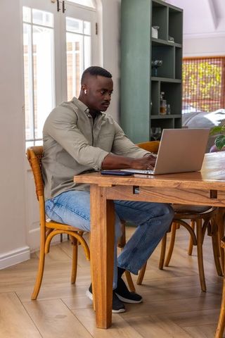 African american man working remotely on laptop at wooden dining table, bright home office