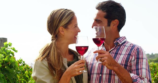 Happy couple smiling and holding wine glasses, enjoying a moment together in a vineyard. The lush grapevines and countryside in the background create a romantic and serene atmosphere. Suitable for use in materials promoting wine, vineyards, romantic getaways, outdoor activities, and relationship celebrations.