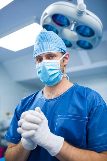 Surgeon in blue scrubs and face mask standing in operating room, preparing for surgery. Ideal for use in healthcare, medical, and hospital-related content, including websites, brochures, and educational materials.