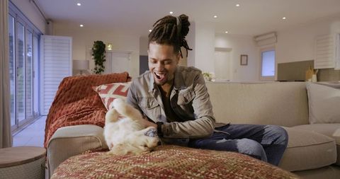 Laughing african american man petting golden retriever on cozy modern living room sofa
