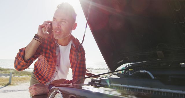 A man, dressed in casual attire with a checkered shirt, is making a call on his phone while standing next to a broken car with the hood up on a sunny day. This image is useful for depicting roadside emergencies, vehicle breakdown scenarios, and the importance of having assistance services. It can be used for automotive repair advertisements, emergency hotline services, and articles related to car maintenance tips.