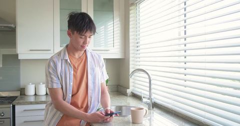 Young man leaning on kitchen counter checking smartphone near ceramic mug in morning light