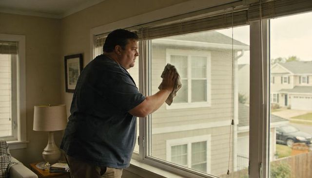Midlife man cleaning living room window with cloth, blinds lifted, suburban home interior routine