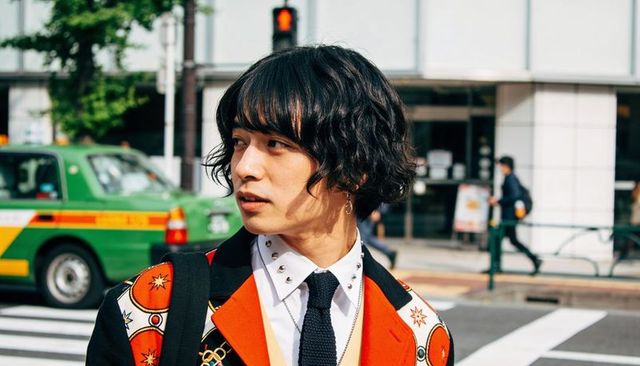 Fashion-forward asian man wearing patterned orange coat crossing urban street with green taxi