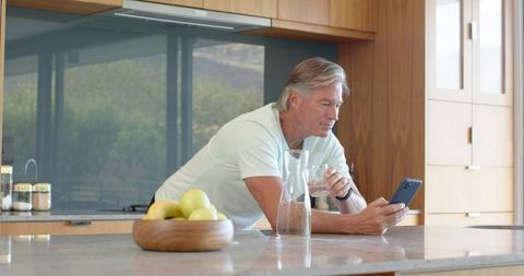 Senior man leaning on modern kitchen island checking smartphone while holding glass