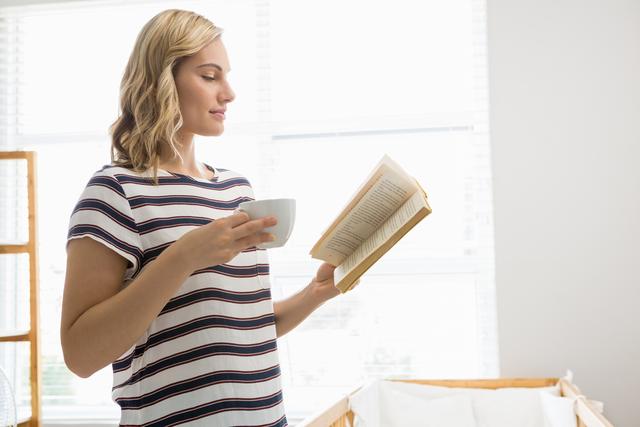 A woman stands near a window with natural light holding a coffee cup and reading a book. She wears a casual striped shirt and appears to be enjoying a peaceful moment at home. This image is perfect for illustrating relaxation, personal time, or promoting home lifestyle products.