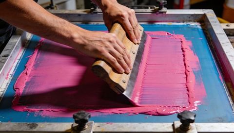 Screen printing magenta ink spreading with wooden squeegee on blue mesh screen closeup