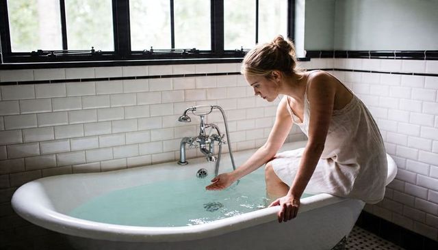 Woman testing water in vintage clawfoot tub beside sunlit window, serene bathroom mood