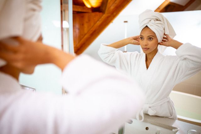 Biracial woman wearing robe and towel turban, looking in mirror. domestic lifestyle and self care, enjoying leisure time at home.