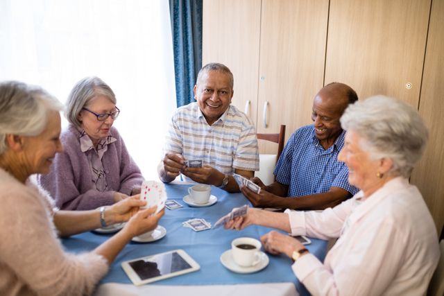 Group of multi-ethnic senior friends playing cards and enjoying coffee at a table in a nursing home. Ideal for use in content related to elderly care, retirement communities, social activities for seniors, and promoting a sense of community and togetherness among the elderly.