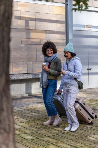 Diverse friends walking with wheeled suitcase and coffee in urban plaza