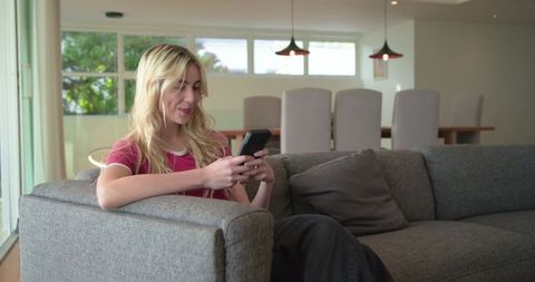 Young woman relaxing on modern sofa using smartphone in bright open-plan living room