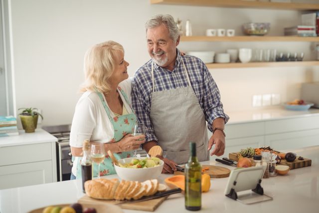 Senior couple enjoying time together while preparing a meal in a modern kitchen. They are smiling and engaging in conversation, showcasing a happy and healthy lifestyle. Ideal for use in advertisements, articles, or blogs related to senior living, healthy eating, family life, and home cooking.