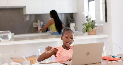 African american daughter studying at kitchen table with laptop while mother cooking