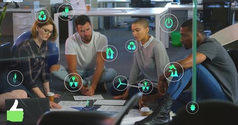 Diverse team reviewing sustainable design plans on office floor with eco technology icons
