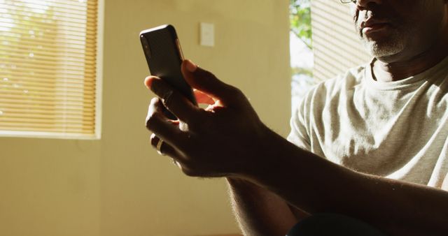 Senior African American man using smartphone in a cozy home setting. Sunlight streaming through window blinds, creating a warm, inviting atmosphere. Man dressed in casual clothing, sitting comfortably. Ideal for use in contexts related to elderly people and technology, home environment, tech-savvy seniors, or personal relaxation.
