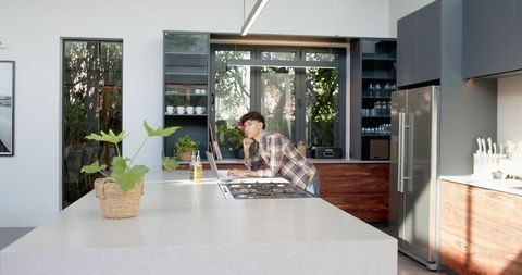 Young Man Multi-tasking with Smartphone and Laptop in Modern Kitchen