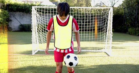 Teen soccer player juggling ball in front of goal wearing red kit and yellow training bib