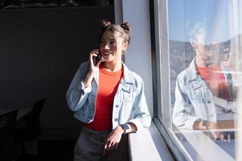 Asian woman talking on smartphone by sunlit window in denim jacket with reflection
