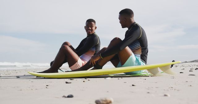 Happy african american teenage twin brothers sitting by surfboards on a beach talking. healthy outdoor family leisure time together.