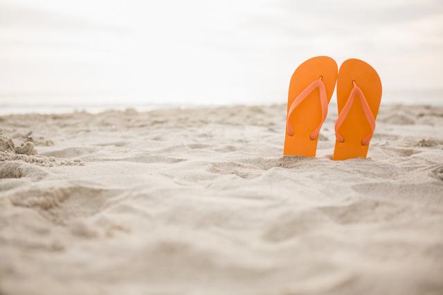 Orange flip flops standing upright in sand on a beach. Ideal for use in travel brochures, summer vacation promotions, beachwear advertisements, and relaxation-themed content.