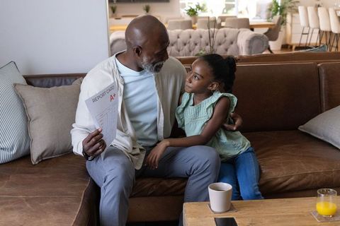 African descent grandfather celebrating granddaughter holding a+ report card on sofa