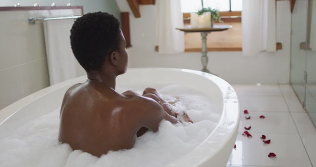 Person soaking in a bubble bath in a sleek, modern bathroom setting with natural light and rose petals on floor conveying self-care and relaxation; ideal for wellness, spa, leisure, home, and lifestyle content.