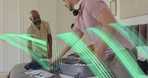 Young man packing suitcase while older man assisting on bed preparing for trip