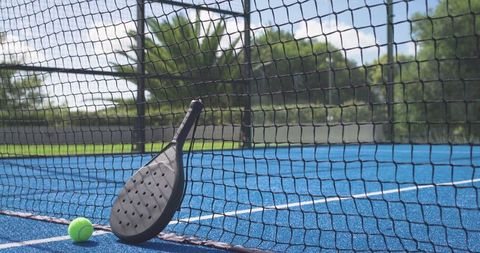Black perforated padel paddle leaning on net with neon tennis ball on vibrant blue court