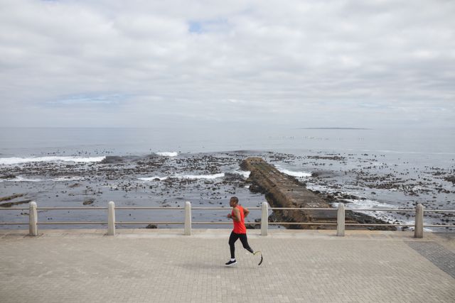 This image captures a disabled biracial man with a prosthetic leg running on a seaside promenade. It is perfect for promoting fitness, healthy lifestyles, and motivation. It can be used in advertisements, articles, and campaigns focused on disability awareness, sports, and outdoor activities.