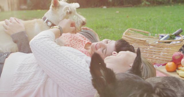 Caucasian lesbian couple playing with their dog while lying on the blanket in garden during picnic. lgbt relationship and lifestyle concept