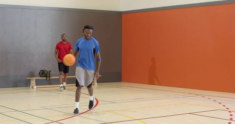 Mid-adult man dribbling basketball on indoor gym court in blue shirt and gray shorts